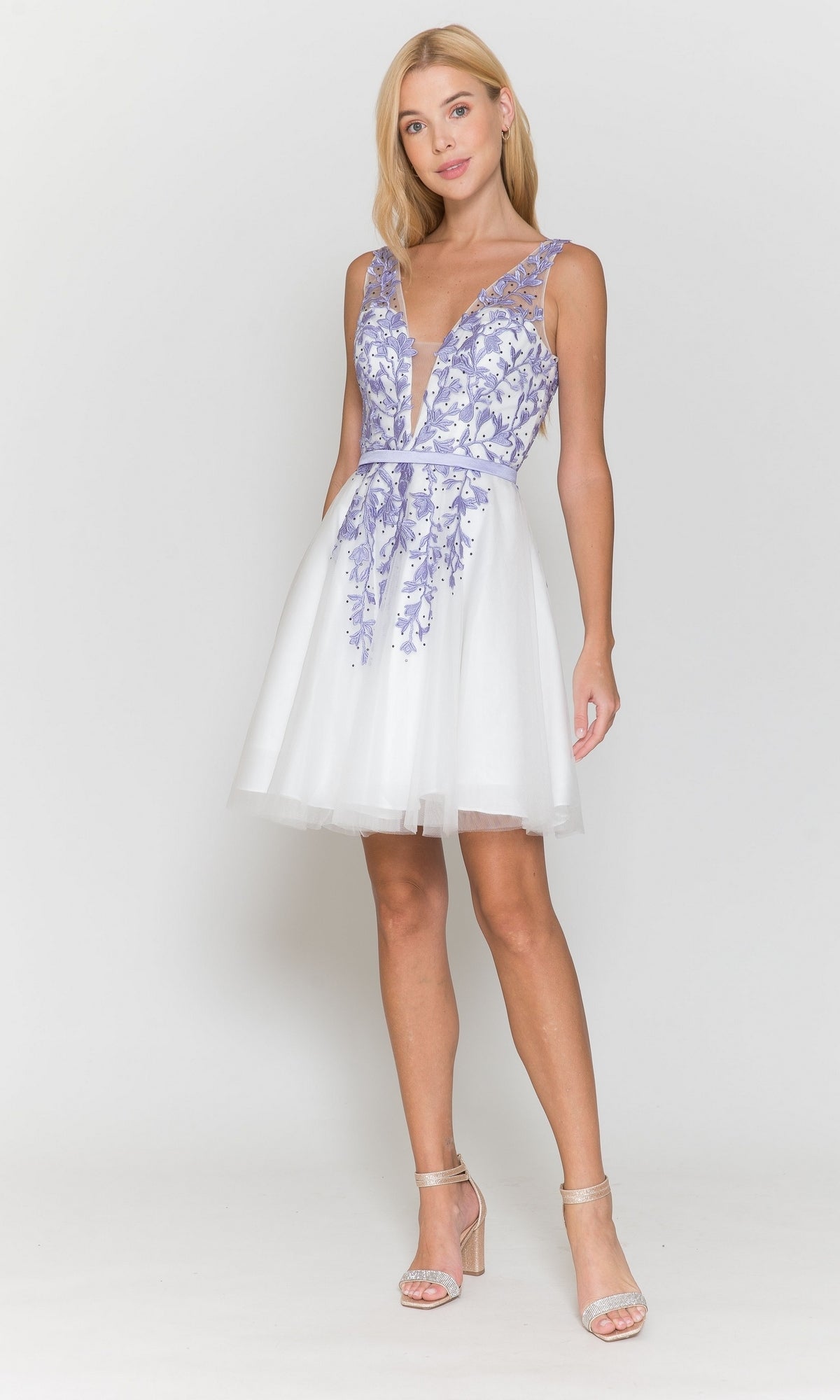 A woman wearing the Tulle and Lace Short A-Line Homecoming Dress 8696, a sleeveless white dress with a deep V neckline and lavender floral embroidery, stands in high heels against a plain white background.